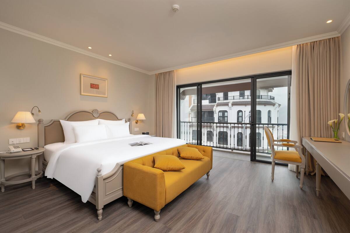 Habitación de hotel con una cama grande, un sofá amarillo, un escritorio con silla y vistas a un edificio a través de un ventanal amplio.