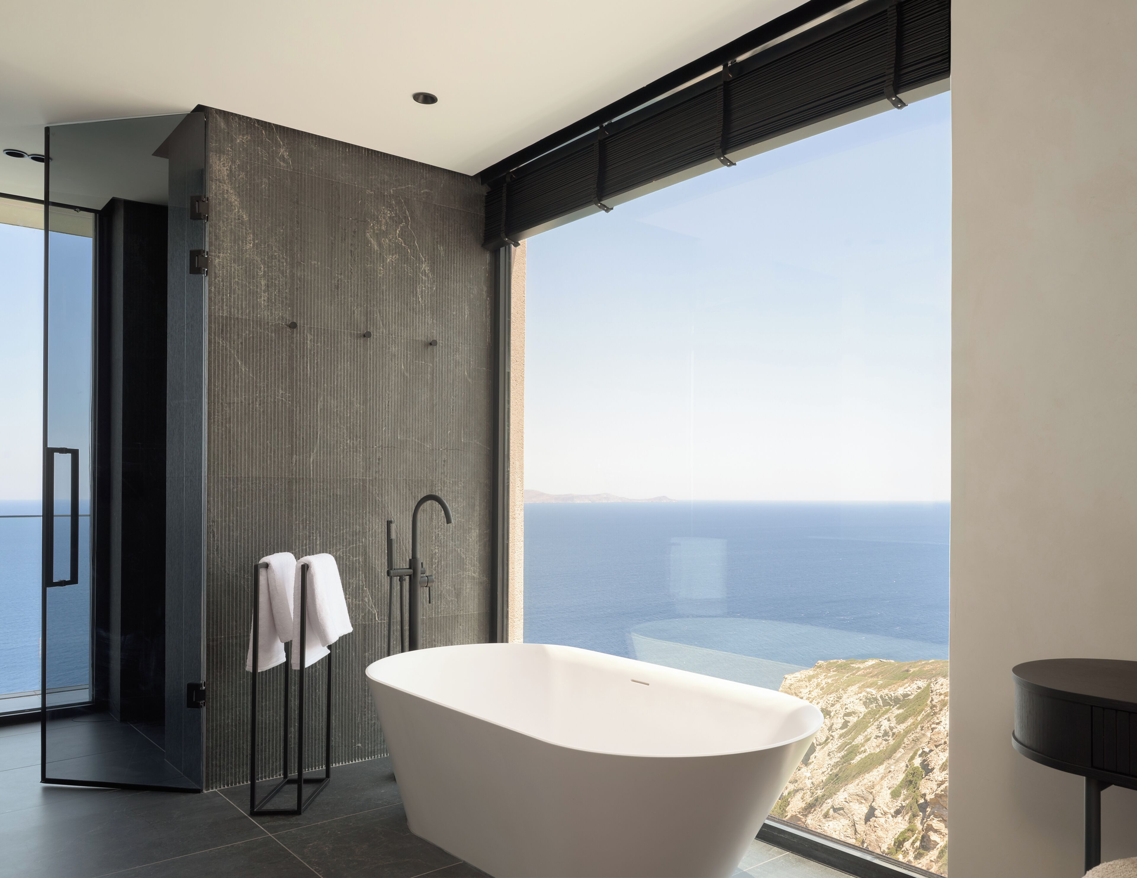 A modern bathroom with a freestanding bathtub, a walk-in shower, and a large window offering a view of the sea.