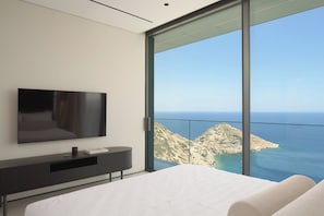 A modern bedroom with a large bed, a flat-screen TV, a dark console table, and a large window offering a view of the sea and rocky terrain. - Seafront Cliff Villa with Infinity Pool & Wellness (Iraklio)
