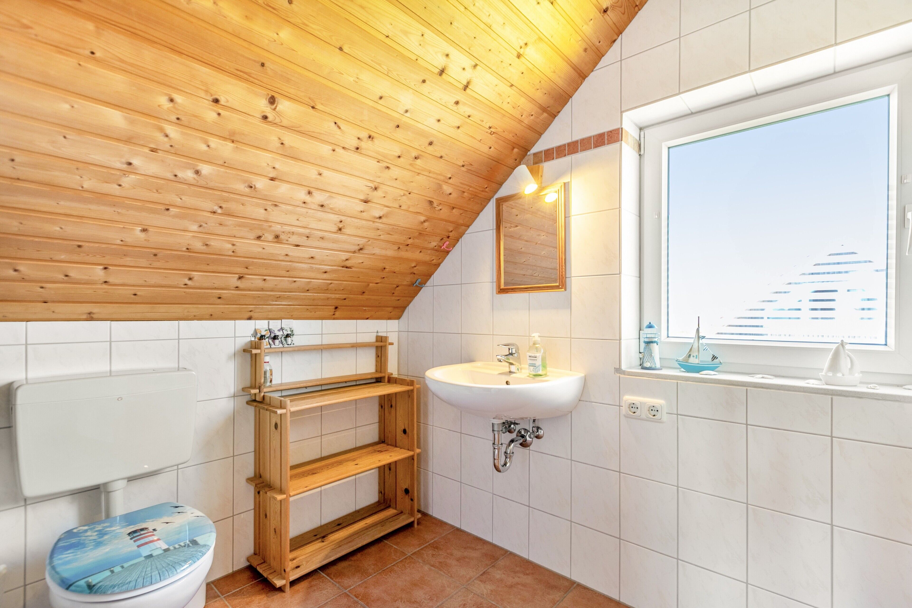 A bathroom with a wooden ceiling, a toilet with a nautical-themed seat, a sink, and a window with a view of a building.