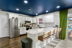A modern kitchen with white cabinets, stainless steel appliances, a central island with bar stools, and a view of a swimming pool through a window. - 5BR, HtdPool, Pickleball, FootPool, 8 min to Beach (Bradenton)