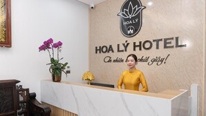 Una recepción con el logotipo y el nombre del Hotel Hoa Ly, un jarrón con flores y una silla de madera.
