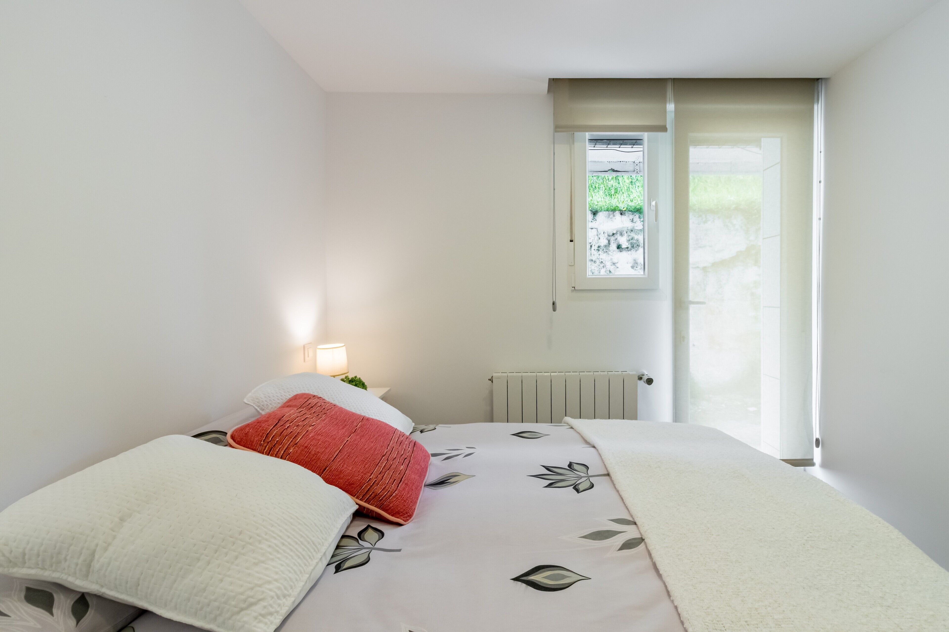 Ein Schlafzimmer mit einem Bett, einem Fenster, einer Heizung und einem Nachttisch mit einer Lampe.