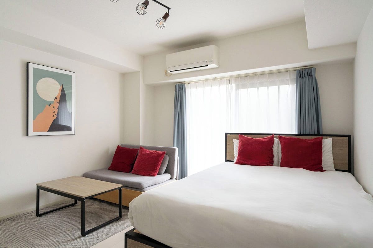 A hotel room with a bed, a sofa, a small table, and an air conditioning unit.