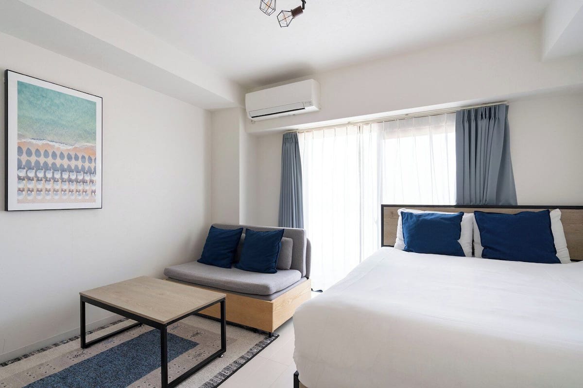 A hotel room with a bed, a sofa, a small table, an air conditioning unit, and a window with curtains.