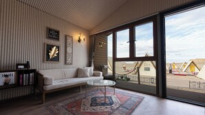 Una sala moderna con un sofá, una mesa de centro de cristal, un tapete decorativo y un ventanal con vistas a una playa y bungalows sobre el agua.