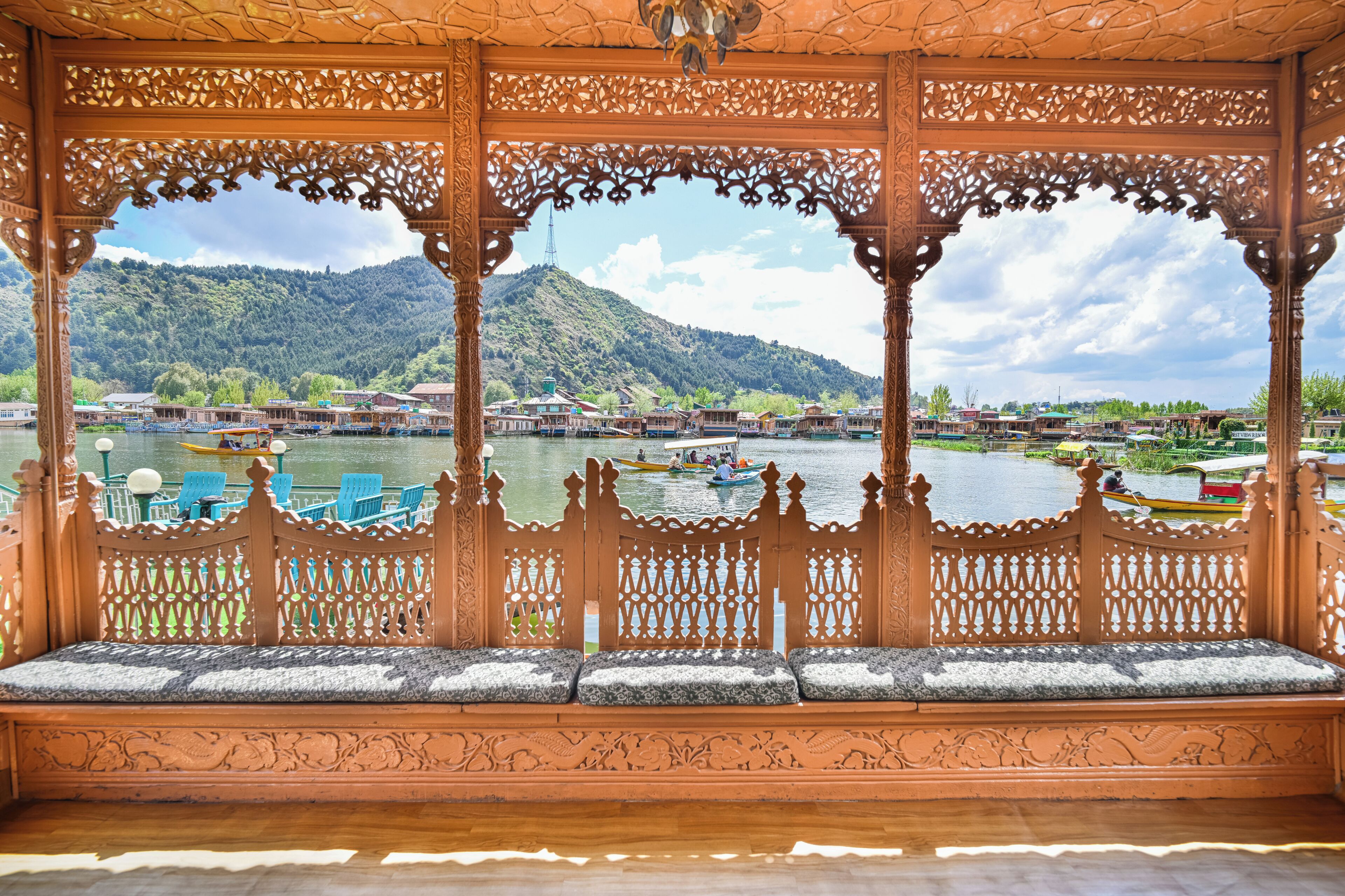 An ornate wooden gazebo with intricate carvings, a bench with cushions, and a view of a river with boats and buildings in the background.