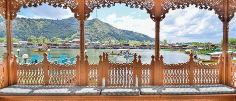 An ornate wooden gazebo with intricate carvings, a bench with cushions, and a view of a river with boats and buildings in the background.
