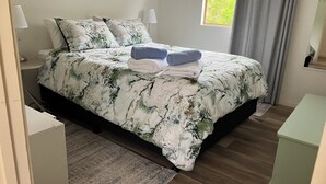 Un dormitorio con cama, almohadas, una mesita de noche, lámpara, ventana con cortinas y vistas al exterior.