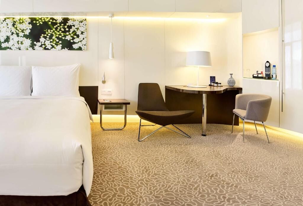 A hotel room with a large bed, two chairs, a small table, a lamp, and a wall-mounted artwork.