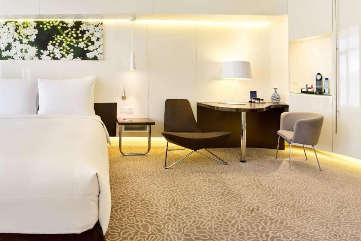 Habitación de hotel con una cama grande, dos sillas, una mesita, una lámpara y un cuadro colgado en la pared.