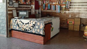 A reception desk with a computer, a reception area with a patterned tablecloth, and shelves stocked with various products. - Chandresh Homestay (Aut)