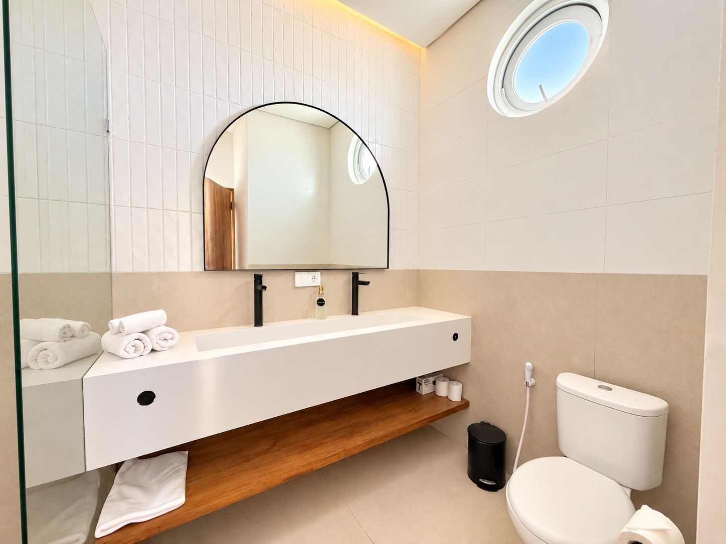 Un baño moderno con un tocador blanco, un espejo redondo, un inodoro y un pequeño cubo de basura.