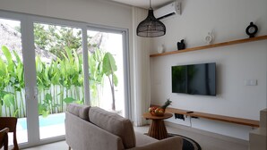 Una sala de estar moderna con un sofá, un televisor montado en la pared, una mesita con fruta y vistas a una piscina y follaje exterior.