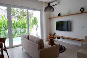 Un salon moderne avec un canapé, un téléviseur fixé au mur, une petite table avec des fruits, et une vue sur une piscine et la verdure à l’extérieur.