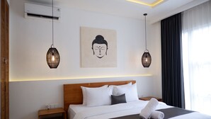 Habitación de hotel con cabecera de madera, ropa de cama blanca, una obra de arte de Buda, dos lámparas colgantes y una mesita de noche con una cesta.