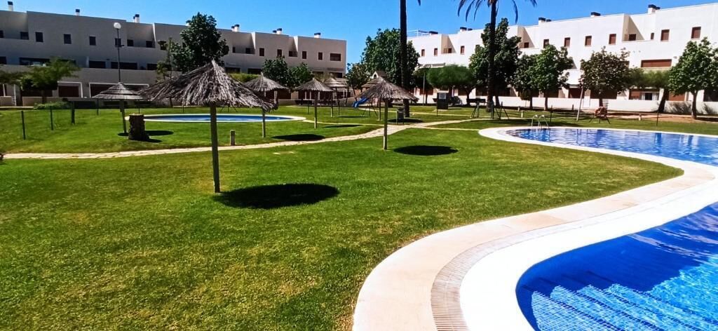 A swimming pool with a curved edge, surrounded by a grassy area with thatched umbrellas, and a building with multiple windows in the background.