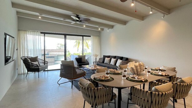 Un vaste espace de vie comprenant un coin repas avec une table pour quatre personnes, entouré de chaises en osier. On y trouve également des ventilateurs de plafond, une grande fenêtre avec des rideaux et un balcon aménagé avec du mobilier d’extérieur.
