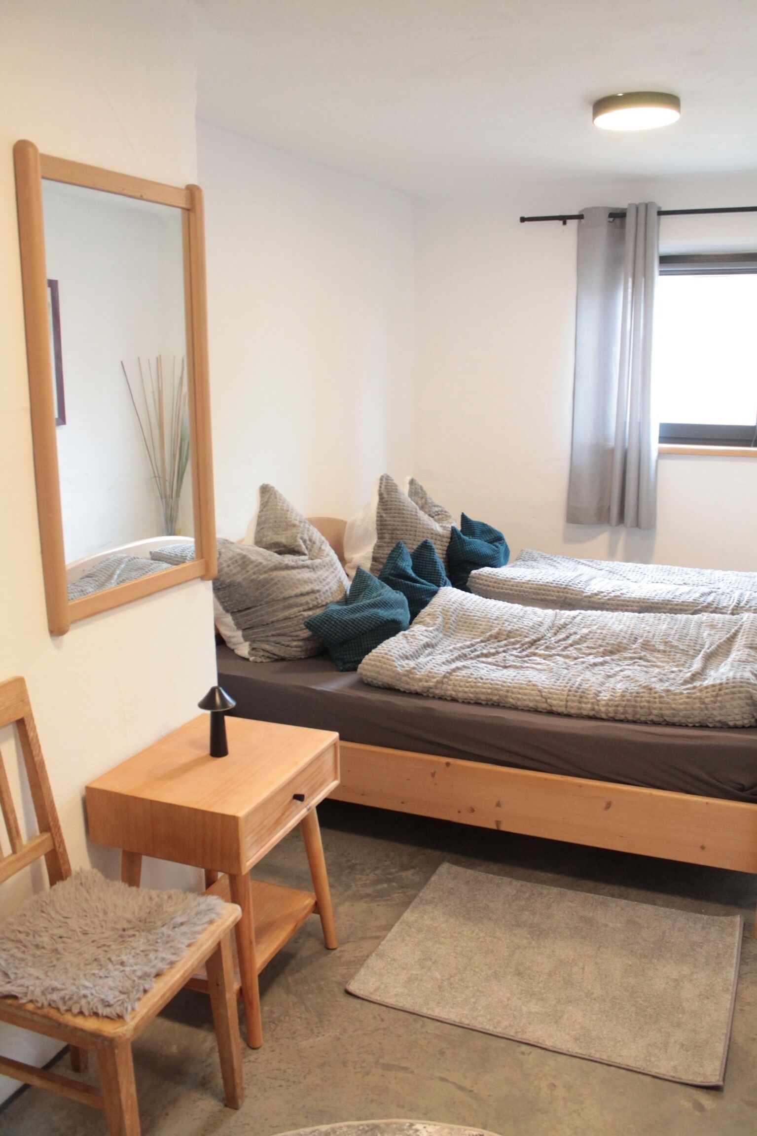 A bedroom with a bed, a desk, a chair, a mirror, and a window with curtains.