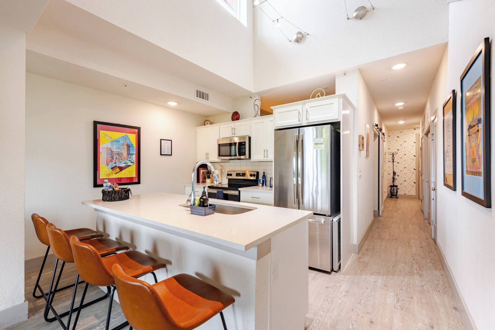 A modern kitchen with a central island, bar stools, stainless steel appliances, and a hallway with patterned wallpaper.