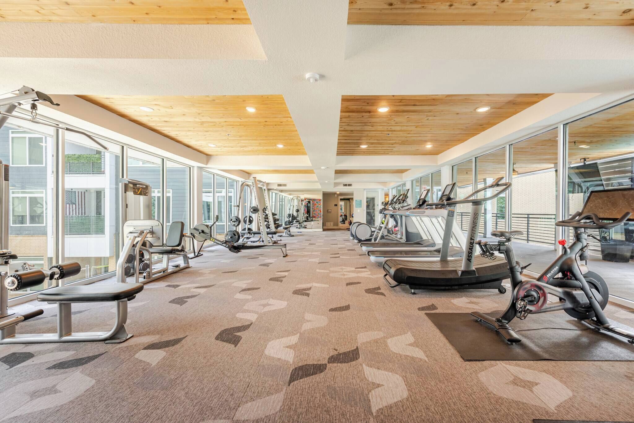 A gym with various exercise equipment, large windows, and a wooden ceiling.