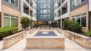 Un immeuble résidentiel à plusieurs étages, doté de balcons, d’une cour intérieure agrémentée d’un élément aquatique et entouré de verdure.