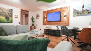 Ein Wohnzimmer mit einer Couch, einem Couchtisch, einem an einer Holzwand montierten Fernseher, einem Essbereich mit Tisch und Stühlen sowie einem Bücherregal.