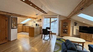 Ein geräumiger Wohnbereich mit hoher Decke, Holzbalken, einem Esstisch, einer Couch und einer Küche mit großem Fenster.