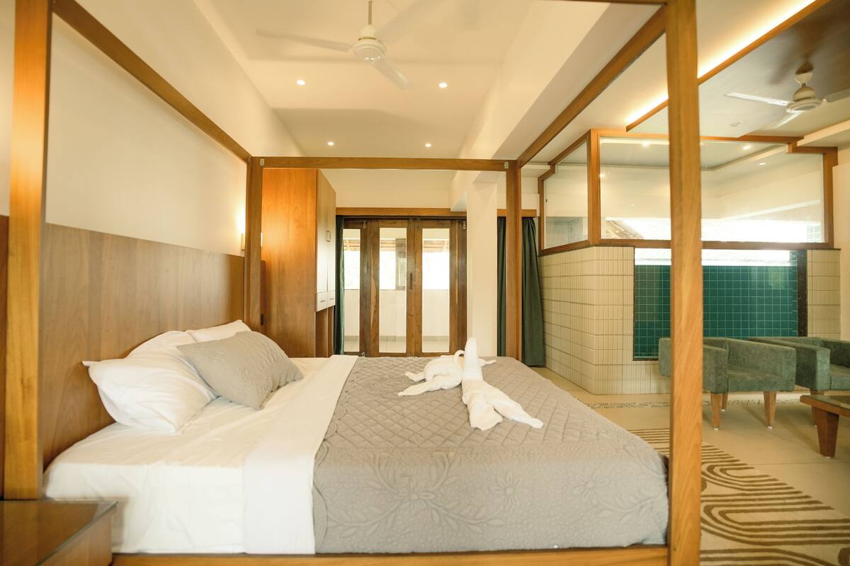 Habitación de hotel con cama, ventilador de techo, baño con pared de azulejos verdes y una zona de estar con una silla y una mesa.