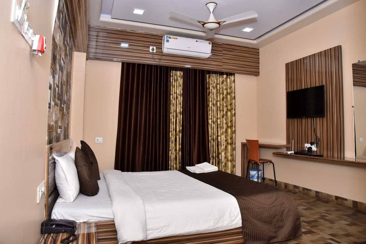 A hotel room with a bed, a flat-screen TV, an air conditioning unit, a ceiling fan, and a window with curtains.
