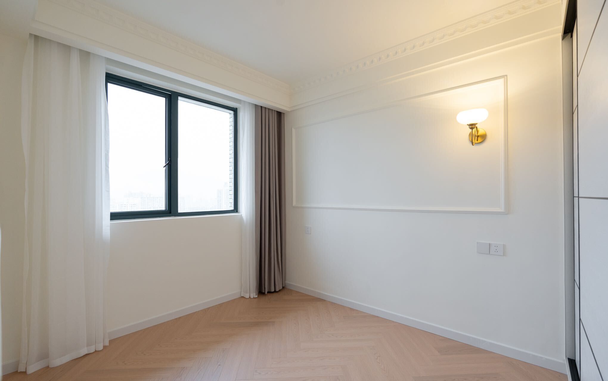 A room with a large window, a wall-mounted light, and a wooden floor.