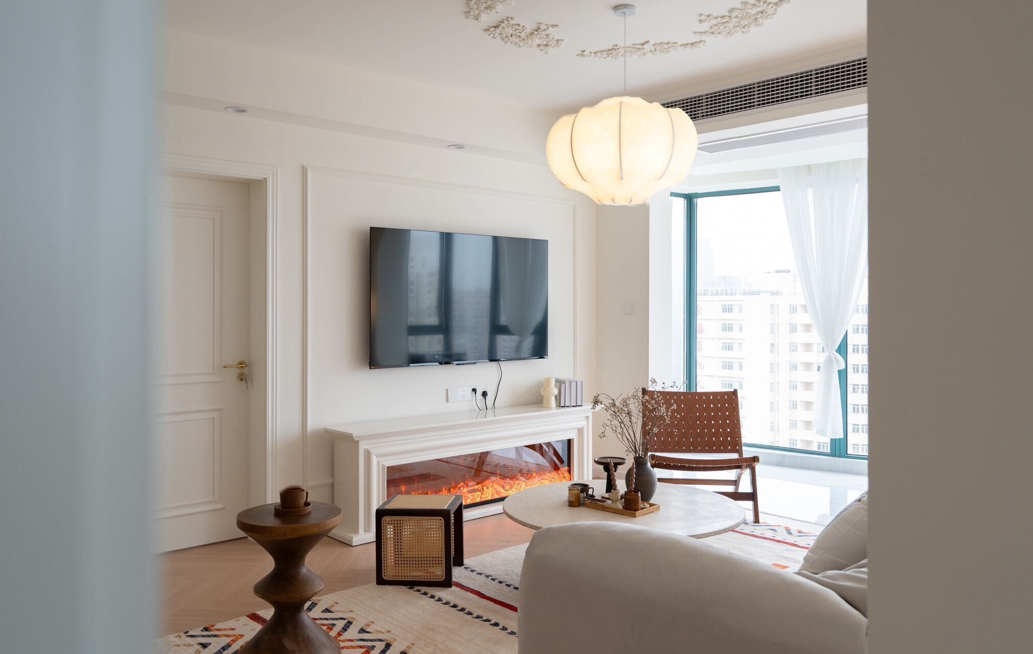 A living room with a fireplace, a television mounted on the wall, a round table, and a large window with a view of buildings.