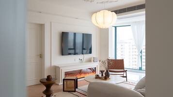 A living room with a fireplace, a television mounted on the wall, a round table, and a large window with a view of buildings.