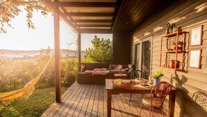 Eine Holzterrasse mit einer Hängematte, einem Sofa, einem Tisch mit einem Stuhl und einem Fenster mit Blick auf Grünfläche.