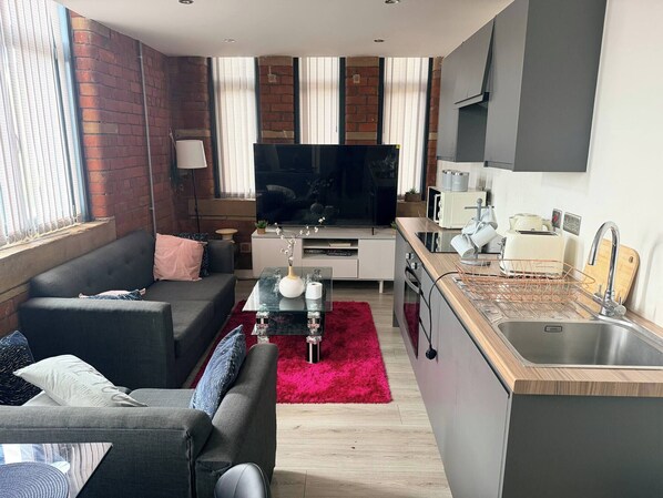 A modern living room with a sofa, TV, and kitchen area including a sink and countertop. - Contractor Friendly 2BR Weekly, Monthly Discount (Bradford)