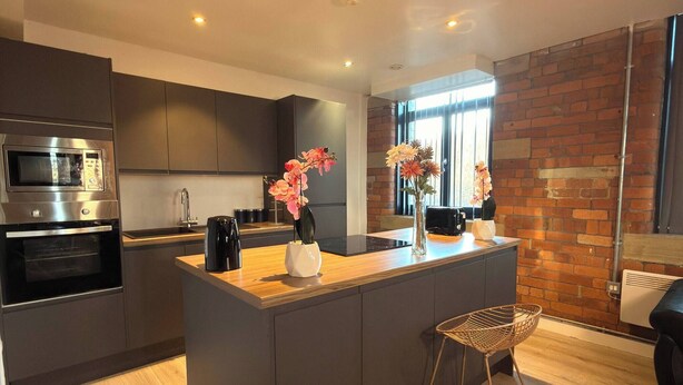A modern kitchen with a central island, dark cabinetry, and a brick accent wall. - Winter stays, Pool, Wi-Fi, Parking, 3double beds (Bradford)