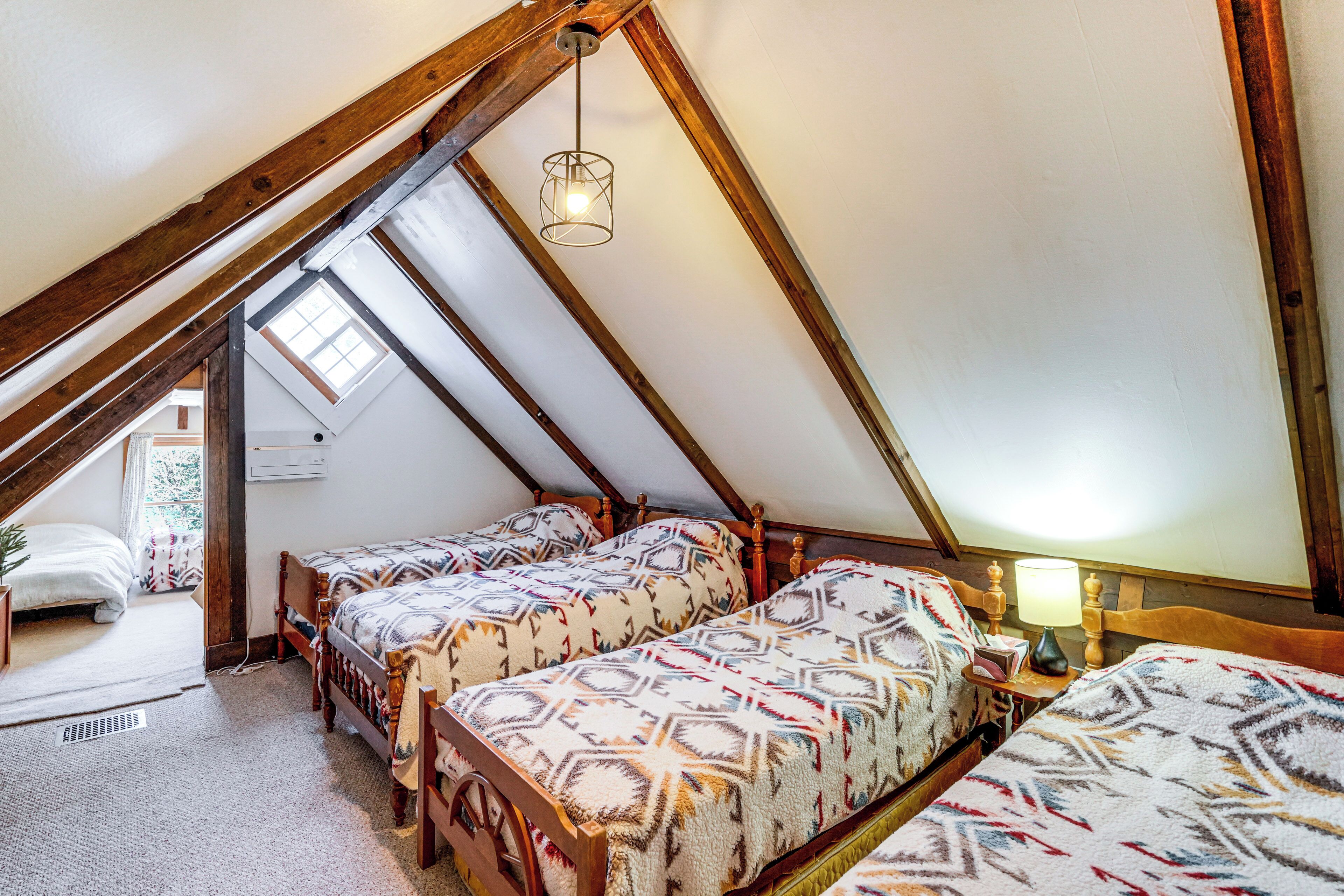 A room with a sloped ceiling, wooden beams, two beds with patterned bedding, a small table with a lamp, and a window.