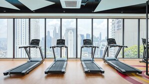 Une salle de sport avec trois tapis de course alignés, de grandes fenêtres donnant sur le paysage urbain et un ventilateur de plafond.