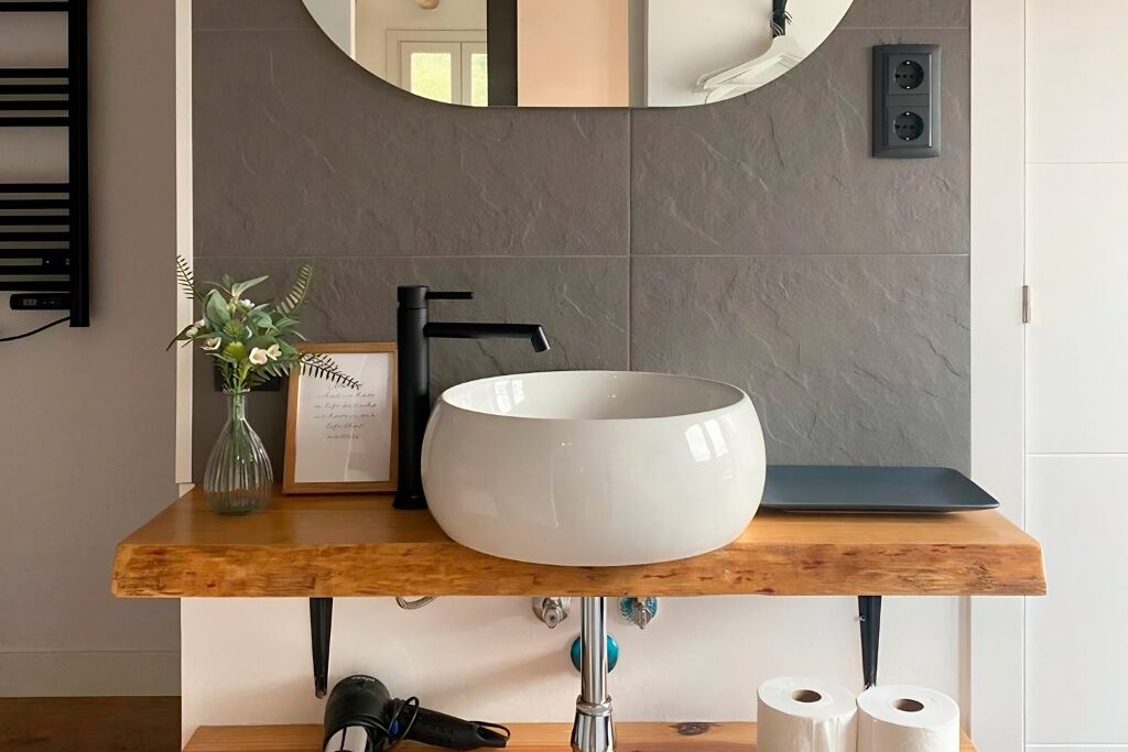 Een badkamer met een ronde witte wasbak, een zwarte kraan, een houten wastafelmeubel, een lijst met een citaat, een vaas met bloemen en een spiegel.