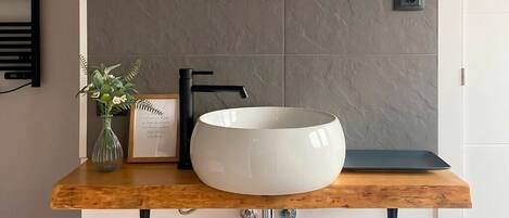 Een badkamer met een ronde witte wasbak, een zwarte kraan, een houten wastafelmeubel, een lijst met een citaat, een vaas met bloemen en een spiegel.