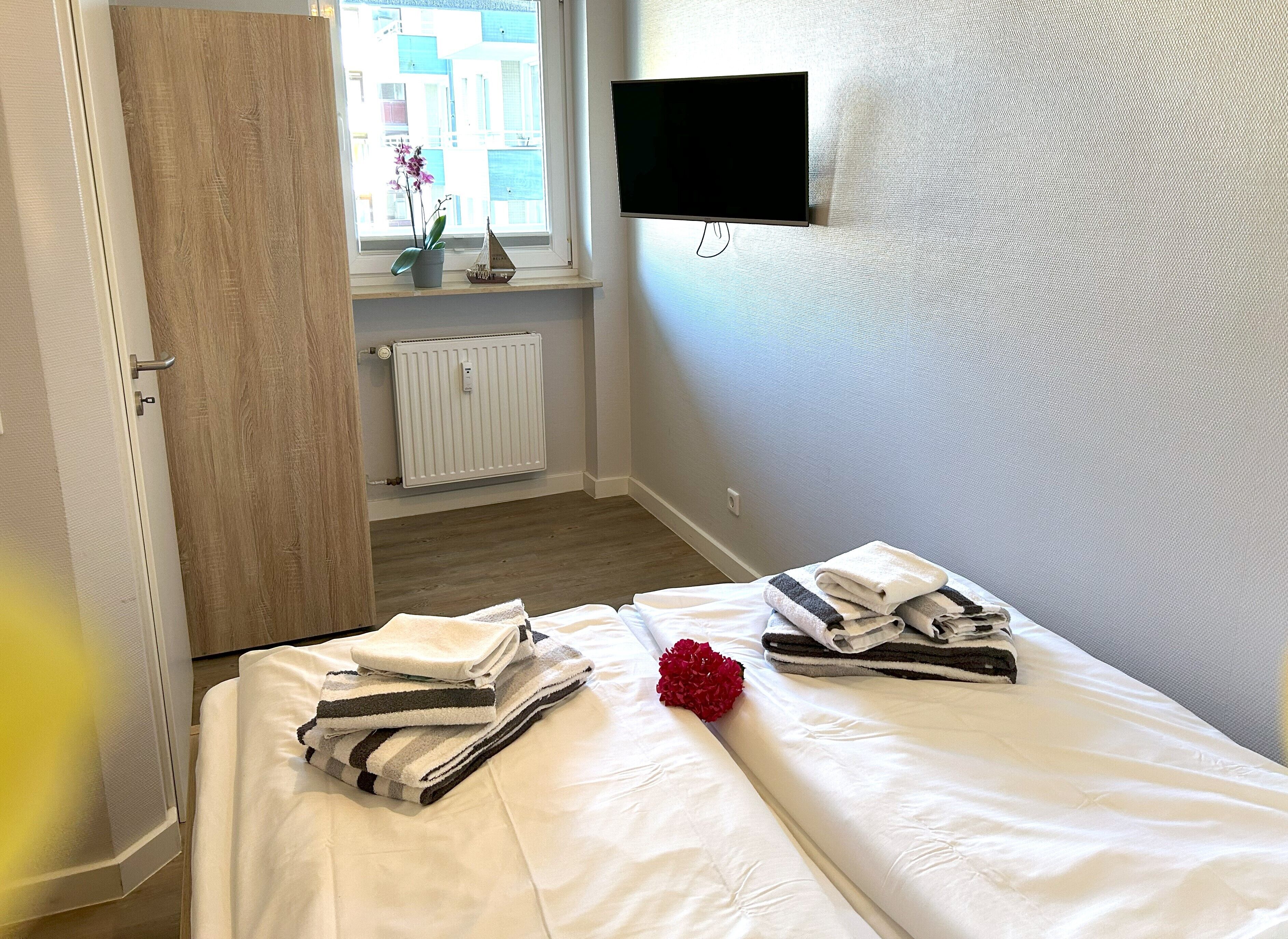 Une chambre d’hôtel avec un lit, un téléviseur fixé au mur, une fenêtre donnant sur des immeubles et un radiateur.