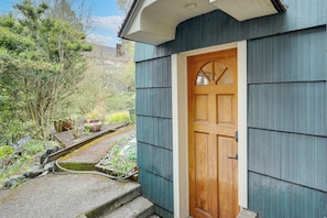 A wooden door with a glass panel, a blue exterior wall, and a garden area with plants and a hose. - Walkable Green Lake Studio w/ Fireplace (Seattle)