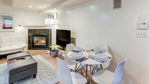 Een moderne woonkamer met een open haard, een flatscreen televisie, een eettafel gedekt voor vier personen en een witte bank.