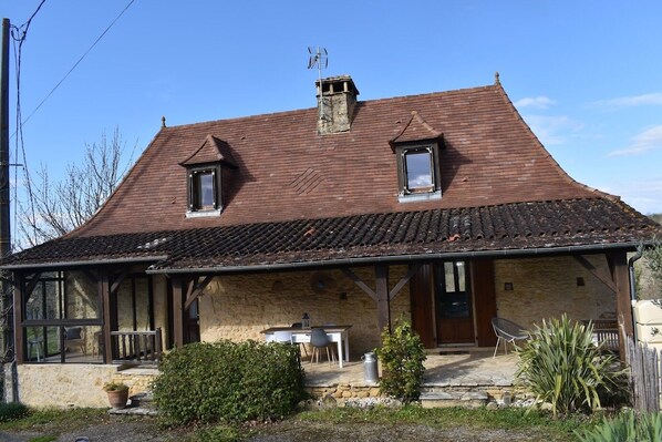 A rustic stone house with a tiled roof, two small windows, a chimney, and a wooden porch with outdoor seating. - Perigordian house in the countryside, calm and nature await you (Orliac)
