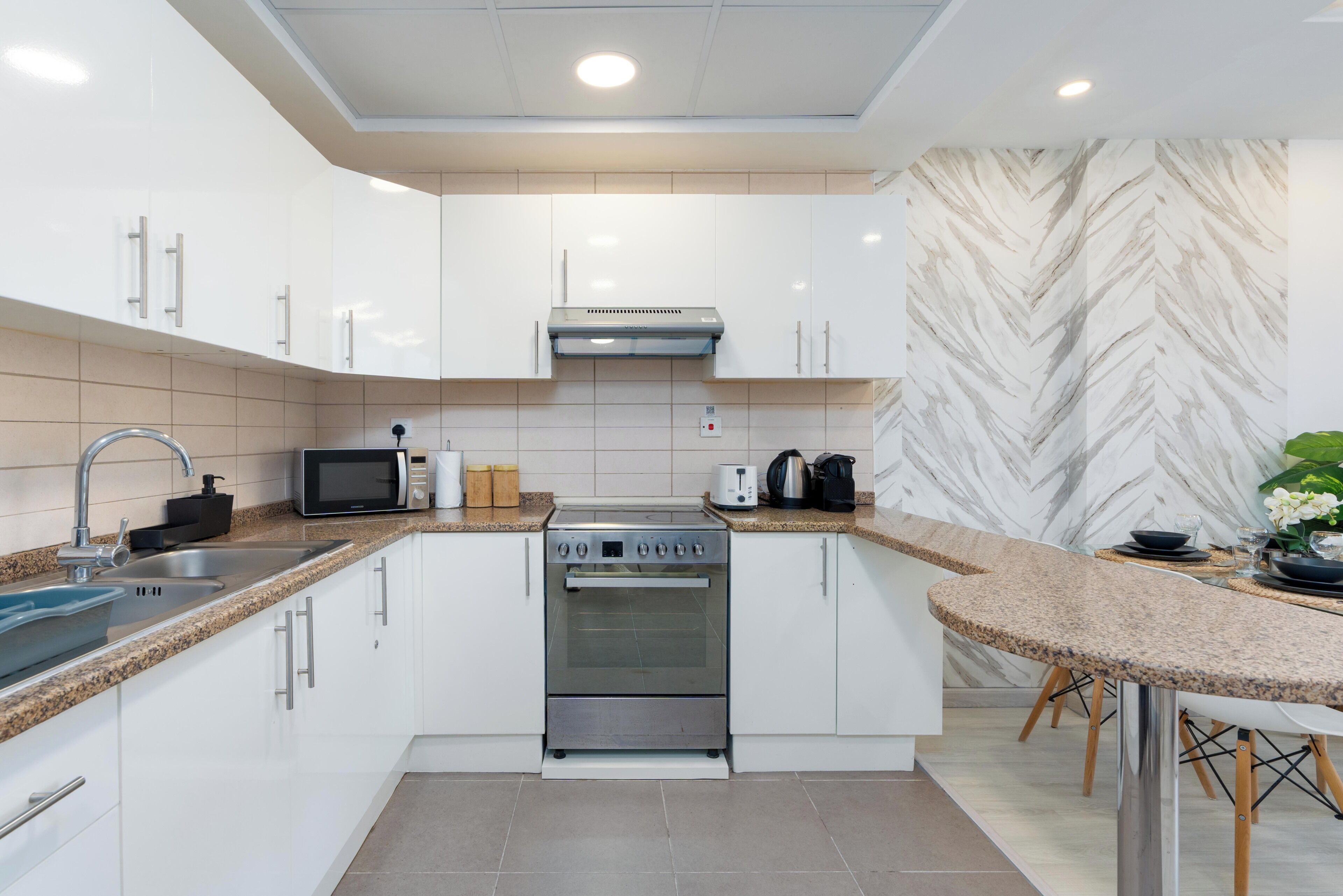 Una cocina moderna con armarios blancos, estufa de acero inoxidable, microondas y un salpicadero con motivo de espiga.