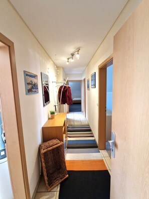 Ein Flur mit einem Holztisch, einem Korbwäscher, Streifen Teppichen und einem Kleiderständer mit einer roten Jacke.