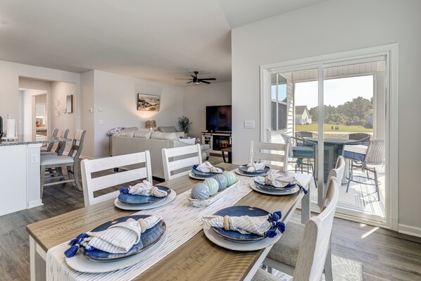 Un comedor con una mesa de madera dispuesta para seis personas, sillas blancas y vistas al exterior a través de amplios ventanales.