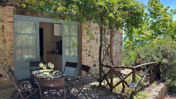 A stone building with a dining area featuring a table and chairs, a kitchen visible through an open door, and a pergola with climbing plants. - Casa Stella Serena by Geco Vacation Rentals (Chiusdino)