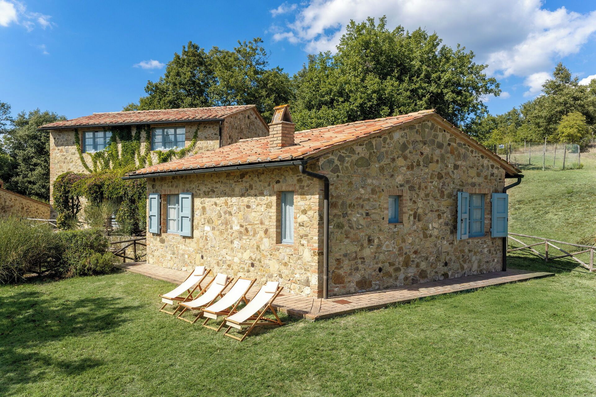 Une maison en pierre avec un toit en tuiles, un petit porche avec des fauteuils de jardin et une cour gazonnée.