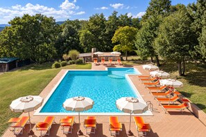 A rectangular swimming pool with a tiled deck, surrounded by lounge chairs and umbrellas, set against a backdrop of trees and a clear sky. - Casa Stella Serena by Geco Vacation Rentals (Chiusdino)